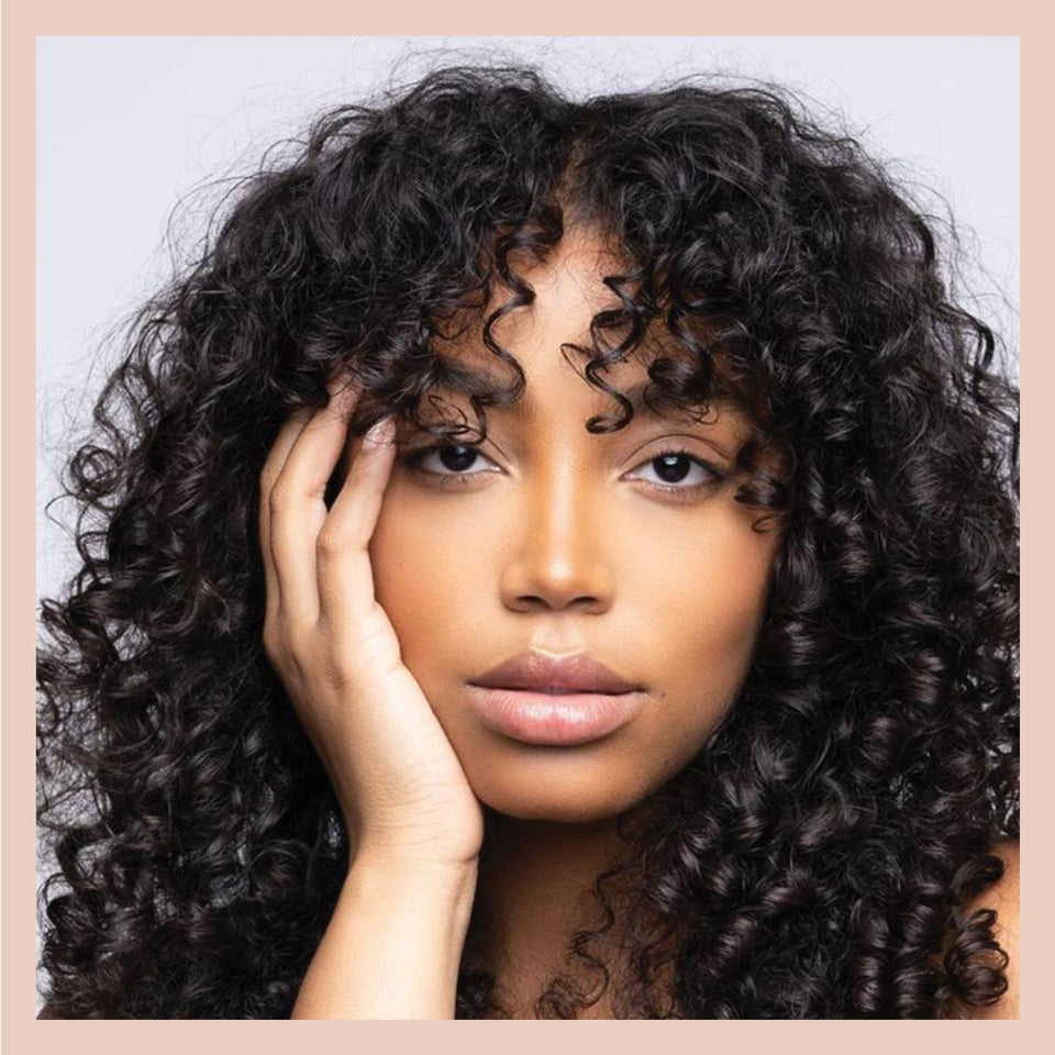 Woman with dark curly hair and curly bangs rests her hand on her face in a close-up beauty portrait