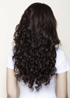 Back view of curly hair extensions showcasing below chest-length, long fuller curls on a person wearing a white shirt