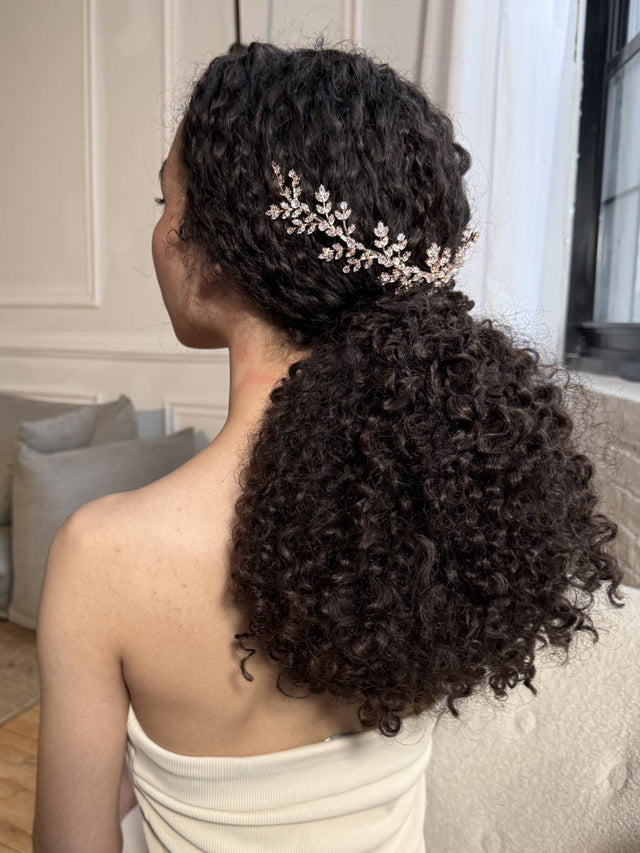 Woman with long curly hair wears a jeweled hair accessory in a low ponytail indoors